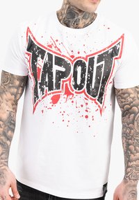 Vyras, vilkintis baltą marškinėlį su dideliu juodu ir raudonu "TAPOUT" logotipu ir raudonais purslais, demonstruojantis tatuiruotas rankas ir kaklą.