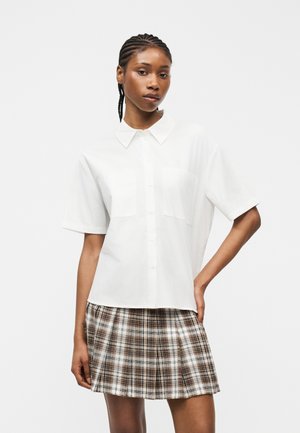 Vrouw met gevlochten haar, gekleed in een wit overhemd met korte mouwen en een bruin geruite geplooide rok, staand met één hand op haar heup.