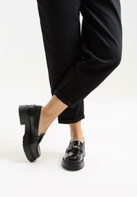 Zwarte gepatineerde leren loafers met een dikke zool, voorzien van een bandaccent over de wreef. Gediend met opgerolde zwarte broeken.