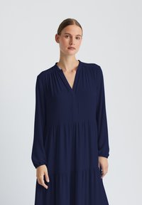 Marineblaues, langärmliges Kleid mit V-Ausschnitt, geraffter Textur an den Schultern und einem gestuften Design. Hergestellt aus leichtem Stoff.