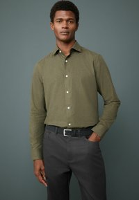 Cămașă verde măsliniu, cu nasturi albi, dintr-un material cu textură subtilă, cu mâneci lungi, purtată cu pantaloni gri închis și o curea neagră.