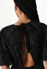 Robe noire avec un tissu texturé à franges et des accents transparents. Détail du dos ouvert maintenu par un nœud en satin. Manches courtes.