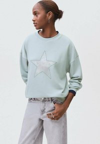 Svetlozeleni pulover z velikim srebrnim zvezdnim vzorcem, okrogel izrez, ohlapen kroj in rebrasti manšeti, v kombinaciji s svetlo sivo denim hlačami.