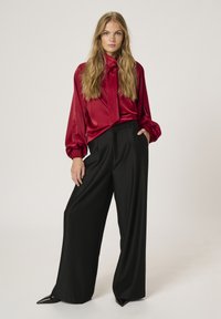 Red satin blouse with a tied neck, long sleeves, and gathered cuffs paired with high-waisted black wide-leg trousers and pointed black heels.