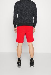 Persona de pie de espaldas vistiendo pantalones cortos rojos, zapatillas deportivas negras con logotipos de Nike visibles y una chaqueta negra con un patrón de líneas onduladas.