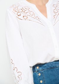 Blusa blanca con intrincados detalles cortados en las mangas y el frente, con un diseño de botones. Combinada con una falda de mezclilla azul con botones dorados.