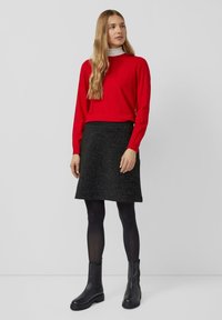 Frau mit langen Haaren, die einen roten Pullover über einem weißen Rollkragenpullover, einem schwarzen Rock, schwarzen Strumpfhosen und schwarzen Ankle Boots trägt, steht vor einem schlichten Hintergrund.