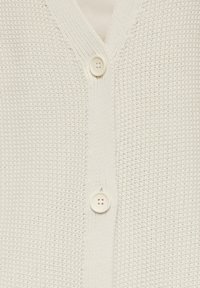 Weiße Strickjacke mit V-Ausschnitt, strukturiertem Stoff und zwei runden Knöpfen vorne. Über einem hellen Oberteil getragen.