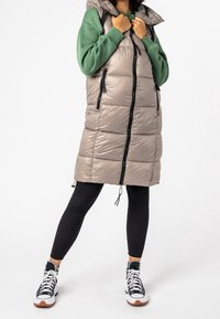 Osoba ubrana w beżową długą kamizelkę puchową na zielonym swetrze, czarnych legginsach oraz czarno-białych butach sportowych na wysokiej podeszwie, stoi na tle białym.