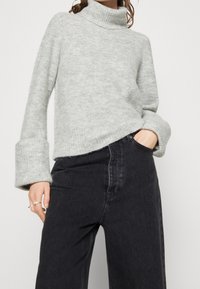 Ljusgrå, grovstickad tröja med hög polokrage och vida ärmar, kombinerad med högmidjade svarta jeans. Mjuk textur och avslappnad passform.