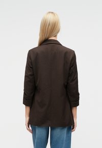 Brązowa marynarka o dopasowanym kroju, z wyciętymi klapami, rękawami do łokcia i gładką strukturą materiału. Posiada pojedynczy szew z tyłu.