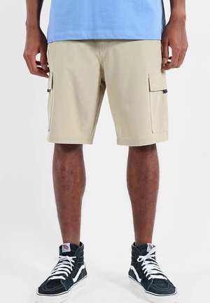 Homme portant un short cargo beige, une chemise bleu clair et des baskets montantes noires avec des lacets blancs, debout devant un fond blanc.