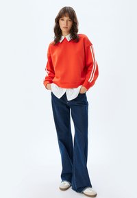 Sudadera roja con rayas blancas en las mangas, superpuesta sobre una camisa blanca con cuello. Combinada con jeans de pierna ancha azules y zapatillas crema.