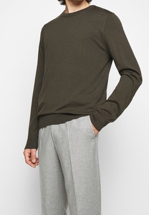 Man wearing an olive green long-sleeve sweater and light grey pleated trousers, standing with one hand in pocket.