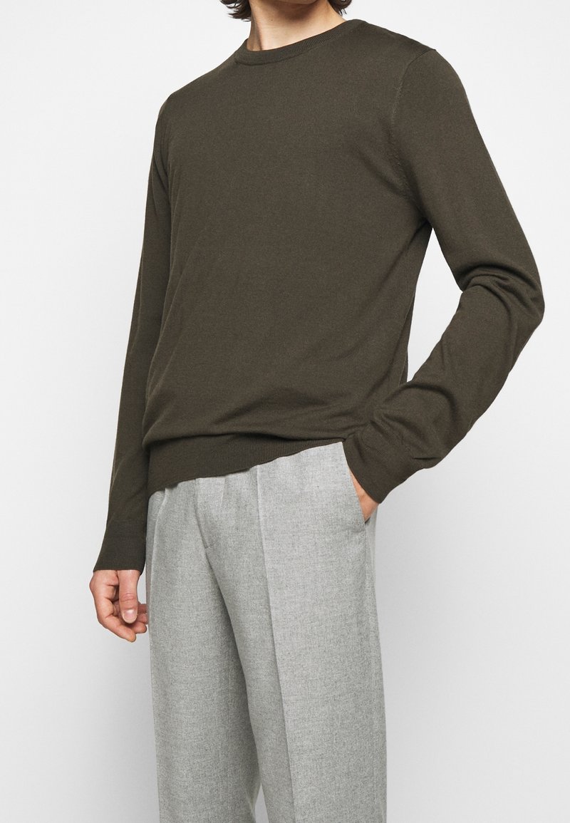 Uomo che indossa un maglione a maniche lunghe verde oliva e pantaloni plissettati grigio chiaro, in piedi con una mano in tasca.