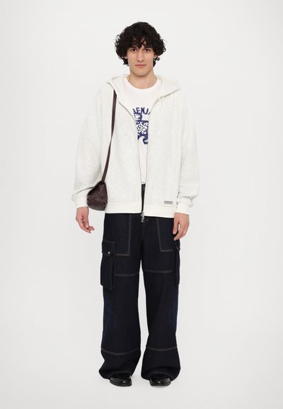 Haut à capuche zippé gris clair avec une finition texturée, superposé à un t-shirt graphique blanc. Pantalons cargo en denim foncé avec poches à rabat et coutures.