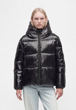 Persona che indossa una giacca imbottita nera lucida con collo alto e zip, abbinata a jeans azzurri, in piedi davanti a uno sfondo semplice.