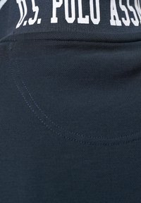 Gros plan d’un tissu bleu foncé avec des coutures cousues et un texte blanc imprimé indiquant "U.S. POLO ASSN" sur une bande texturée.