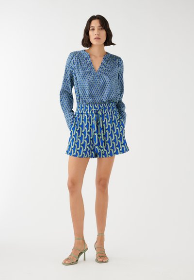 Femme debout portant un chemisier bleu à manches longues à motifs et un short taille haute assorti avec des talons verts à brides sur fond blanc.