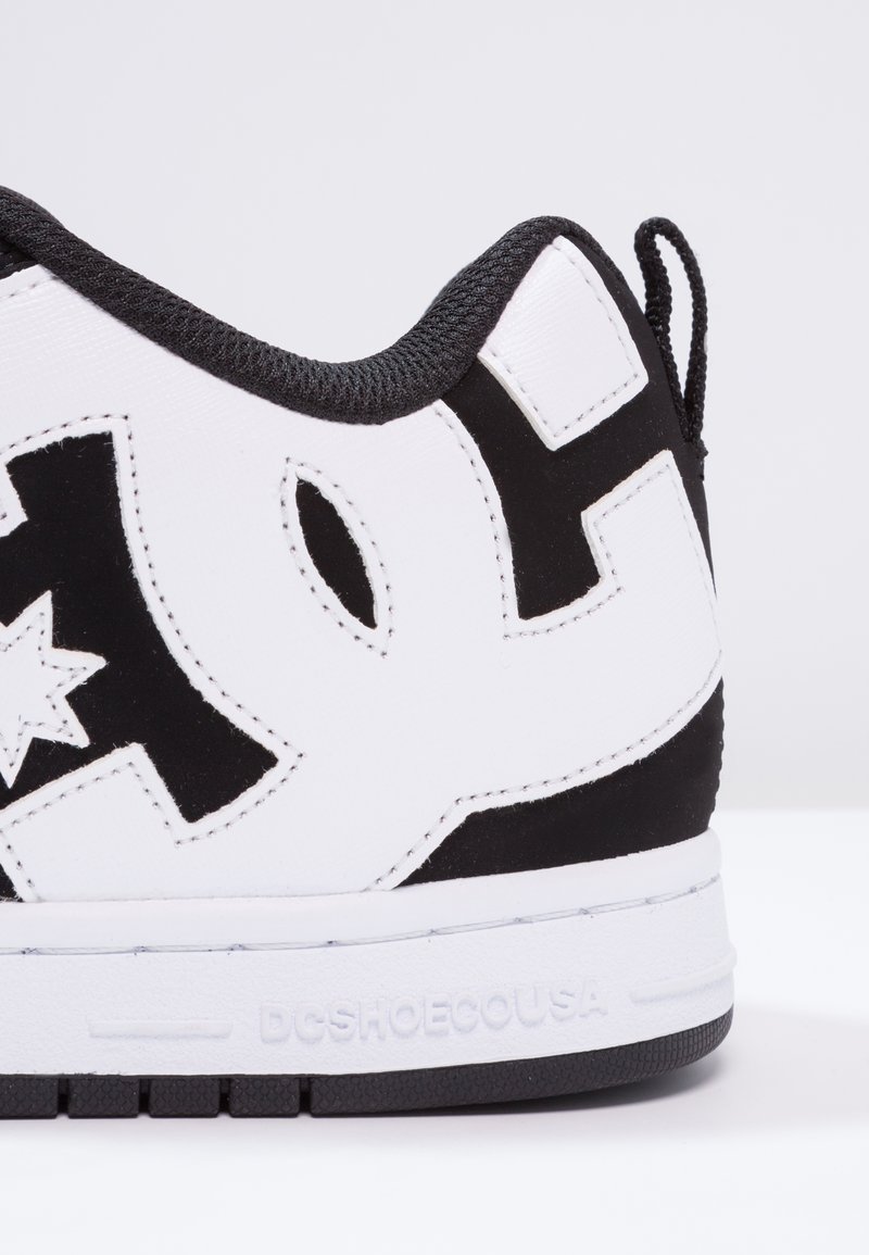 Zapatilla blanca con acentos negros, que presenta materiales lisos y texturizados, un gran logo, detalles de costura y suela de goma negra.