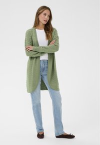 Zelena pletena jopa z vzorcem teksture, odprtim sprednjim delom in dolgimi rokavi, kombinirana s svetlo modrimi jeans hlačami in rjavimi balerinkami.