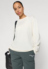 Long-sleeve cream cotton shirt with a crew neckline, paired with dark green sweatpants featuring white logo text on the thigh.