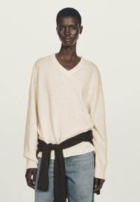 Beige Kaschmirpullover mit V-Ausschnitt und gerippten Bündchen, kombiniert mit blauen Jeans und einem dunkelbraunen Strickgürtel, der in der Taille gebunden ist.