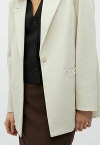 Donna che indossa un blazer color crema sopra una camicia nera con bottoni e una gonna marrone a coste, con la mano rilassata al fianco.