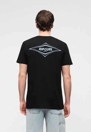 Mężczyzna zwrócony tyłem, ubrany w czarną koszulkę Rip Curl z niebieskim logo oraz jasnoniebieskie dżinsy, na jednolitym jasnym tle.