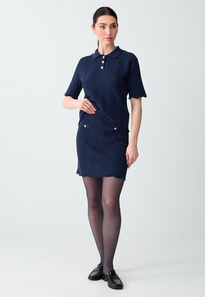 Kobieta ubrana w granatową teksturowaną sukienkę polo z krótkim rękawem i złotymi guzikami, czarne przezroczyste rajstopy oraz czarne mokasyny, stojąca na jednolitym tle.