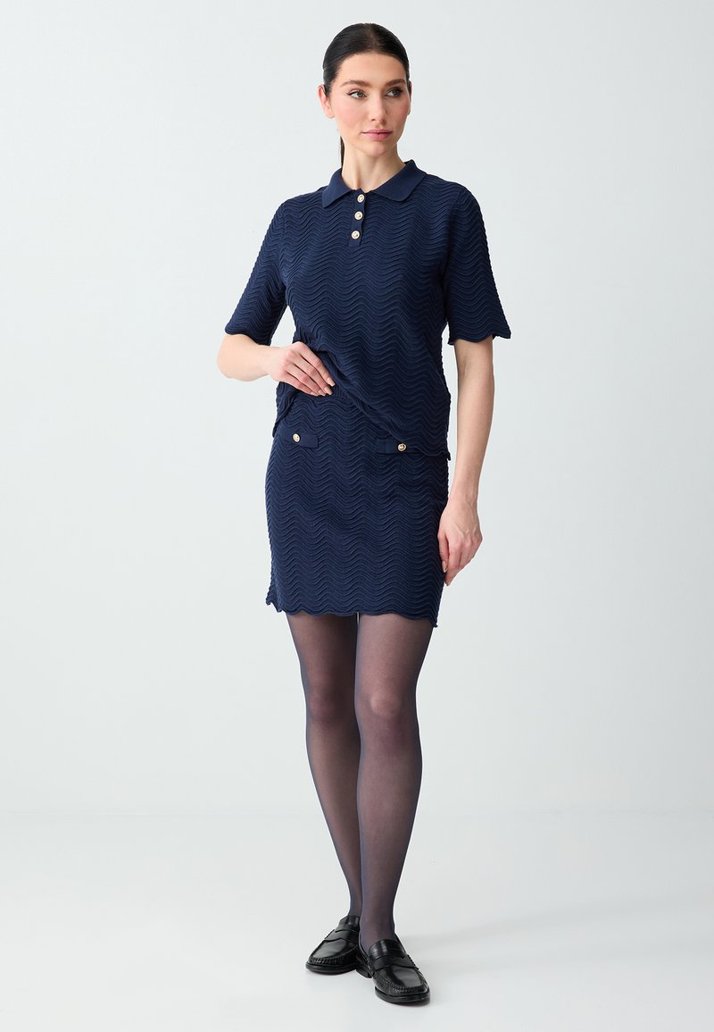 Femme portant une robe polo courte texturée bleu marine avec des boutons dorés, des collants noirs transparents et des mocassins noirs, debout devant un fond uni.