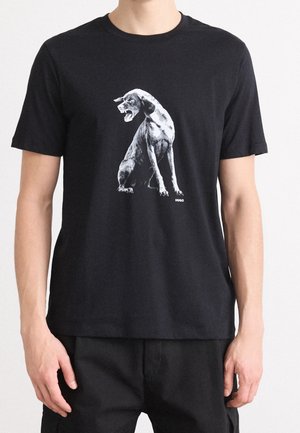 Schwarzes T-Shirt mit einer realistischen Graustufengrafik eines knurrenden Hundes, der sitzt, getragen von einer Person mit schwarzer Hose vor weißem Hintergrund.