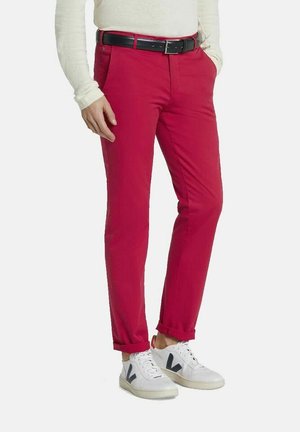 Mann trägt leuchtend rote Slim-Fit-Hose mit schwarzem Gürtel und weißen Turnschuhen, steht mit einer Hand in der Tasche.