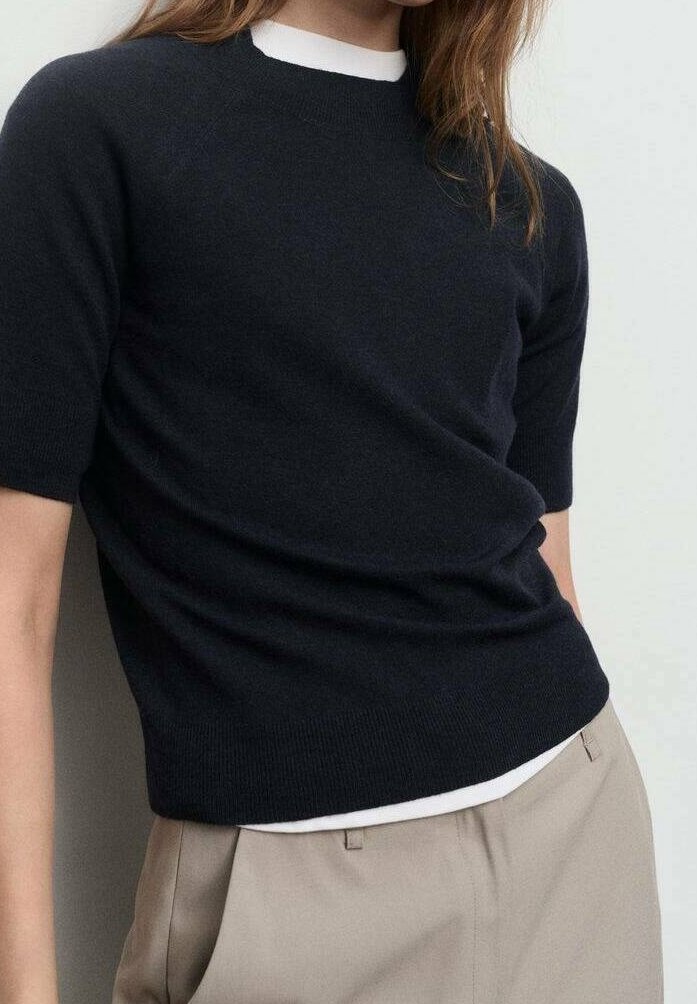 Buste d'une personne portant un pull noir ajusté à manches courtes sur un sous-vêtement blanc, associé à un pantalon beige taille haute.