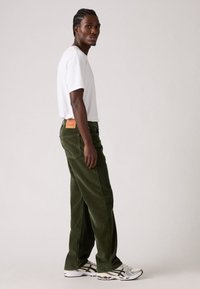 Jonge man met gevlochten haar, gekleed in een wit T-shirt, olijfgroene corduroy broek en witte hardloopschoenen, staande tegen een effen achtergrond.
