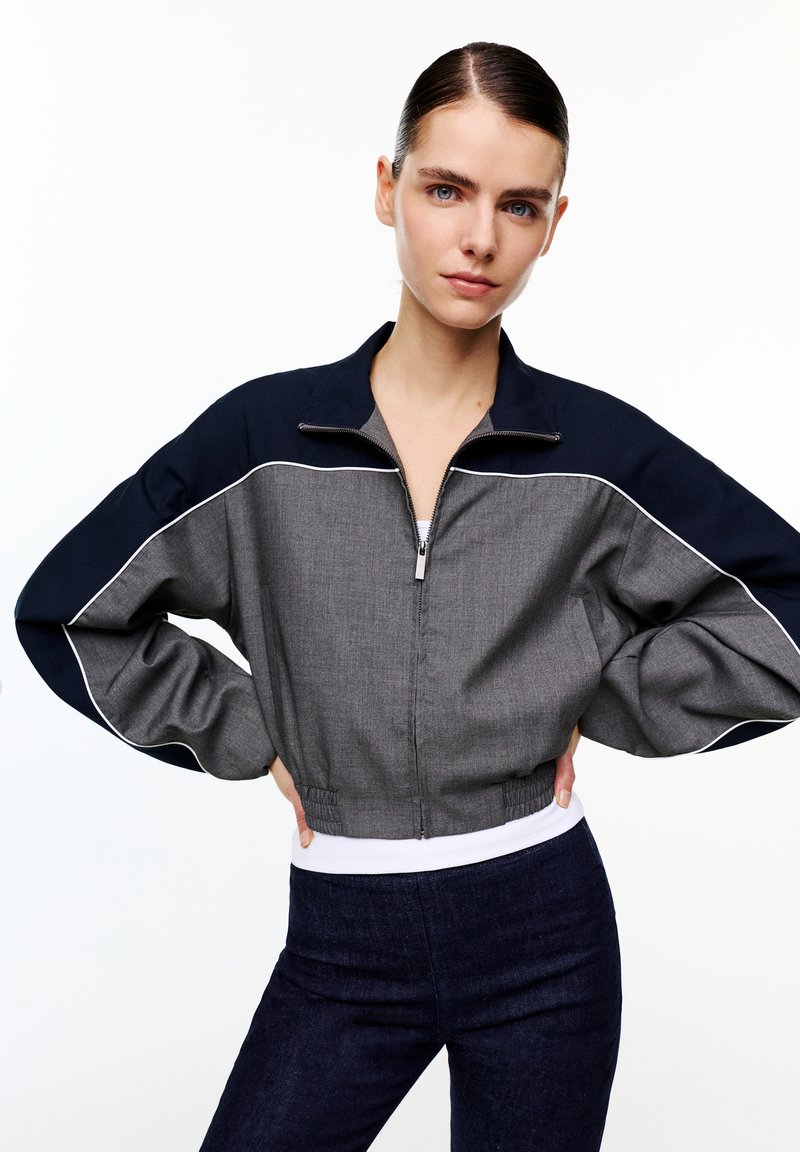 Femme aux cheveux foncés coiffés en chignon élégant, portant une veste zippée grise et noire avec un liseré blanc, debout les mains sur les hanches sur un fond blanc.