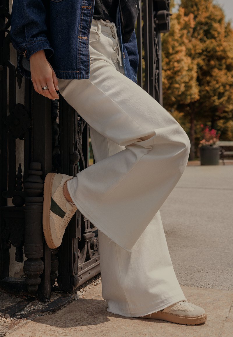 Kremowe spodnie w szerokim kroju, wykonane z gładkiego materiału, w połączeniu z beżowymi trampkami z zielonymi akcentami. Na górze widoczna dżinsowa kurtka.