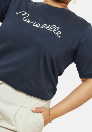 Marineblaues Baumwoll-T-Shirt mit weißer, kursiver Aufschrift "Marseille", Rundhalsausschnitt, kurze Ärmel, kombiniert mit beigen Hosen.