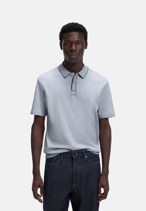 Hombre con camisa tipo polo de manga corta gris claro con detalles oscuros y vaqueros azul oscuro, de pie frente a un fondo blanco liso.