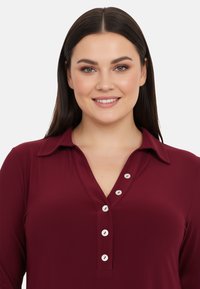 Chemise bordeaux à col avec une patte de boutonnage dotée de boutons blancs, fabriquée dans un tissu lisse.