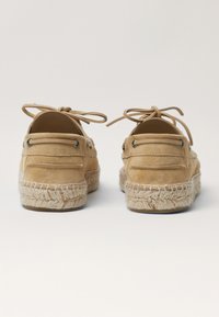 Sandfarbene Wildleder-Espadrilles mit einer gewebten Jutsohle. Mit Lederbindern und Metallösen ausgestattet. Abgerundete Ferse und glatte Textur.