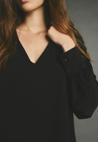 Čern�á blůza s výstřihem do V, dlouhými rukávy se sklady a knoflíkovými detaily na manžetách. Hladká textilie.