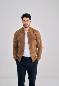 Ruskea mokkanahkainen bombertakki, jossa on resoriset hihansuut ja kaulus, vetoketjulliset taskut, päällä valkoinen t-paita ja yhdessä tummien housujen kanssa.