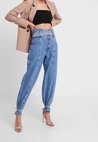 Pantalones vaqueros azules de talle alto con pierna ancha, cierre de botones y puños anudados. Combinados con un top corto negro y una blazer beige.