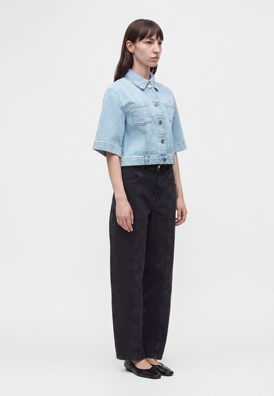 Femme debout portant une veste en jean courte bleu clair, un jean noir ample taille haute et des ballerines noires pointues sur un fond uni.