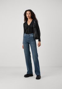 Mulher com cabelo encaracolado, vestindo uma blusa preta pregueada com botões, jeans de cintura alta azuis, sapatos pretos robustos e a carregar uma bolsa preta de ombro.