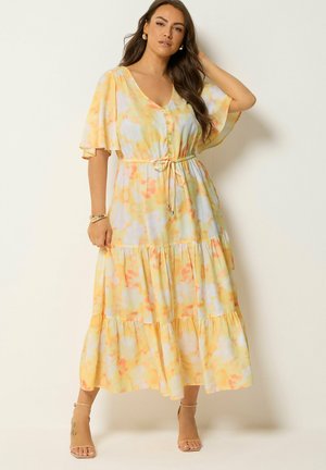 Frau mit langen braunen Haaren, die ein gelbes und orangefarbenes Midikleid mit Blumenmuster, flatternden Ärmeln und Taillenschnürung trägt, posiert mit einer Hand im Haar.