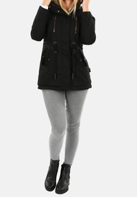 Parka noire avec capuche, fermeture éclair avant et poches à boutons, taille à cordon, et tissu texturé, associée à un jean skinny gris et des bottes noires.