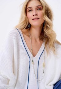 Witte gebreide pullover met blauwe accenten, met een diepe V-hals en een losse pasvorm. Gecombineerd met een gelaagde kralenketting.