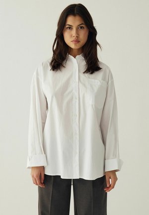 Jeune femme aux cheveux foncés porte une chemise blanche oversize boutonnée et un pantalon foncé, debout devant un fond clair uni.